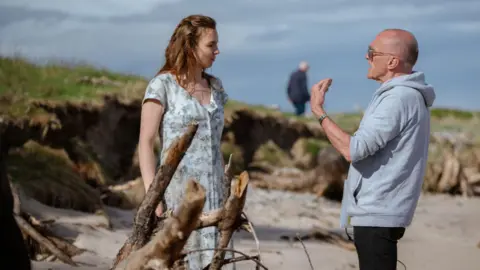 Sony Images Jodie Comer wears a long floral dress as she stands on a beach on the set of 28 Years Later talking to Danny Boyle - he has a shaved head and wears sunglasses and a pale grey hoody. He is gesturing with his hand as he speaks to the actress. Washed-up pieces of wood can be seen in the foreground and there are grassy sandbanks behind them 