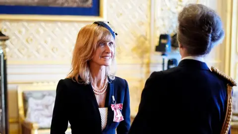 PA Media Jackie Bird smiles at the Princess Royal after receiving her MBE from the Princess Royal which is displayed on her lapel, attached to a pink ribbon in the shape of a bow. Jackie is wearing a dark blue or black suit with a white top underneath. She has a string of pearls around her neck. You can only see the back of the Princess Royal