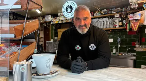 Martin Heath/BBC Liberato Lionetti with short white hair and beard, wearing a black top with two circular logos on it, and grey gloves, stands behind the counter of a coffee shop. There are packs of coffee behind him on wooden shelves, a wooden display shelf to his right and a grey coffee cup in the foreground.