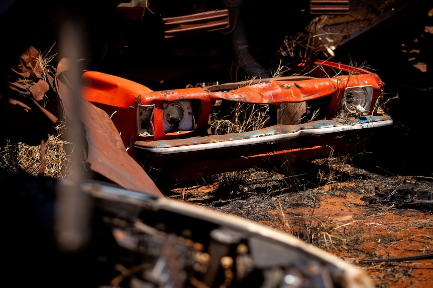 Front of a wrecked orange car sits surrounded by rusted parts of cars around it. 