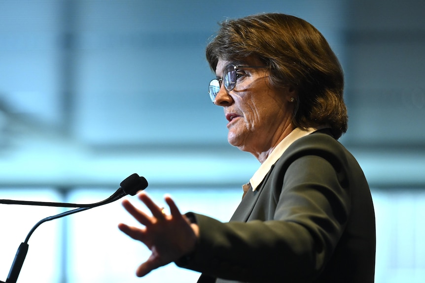 Michelle Bullock, working a suit and glasses, gestures with her hands while speaking in front of a lectern.