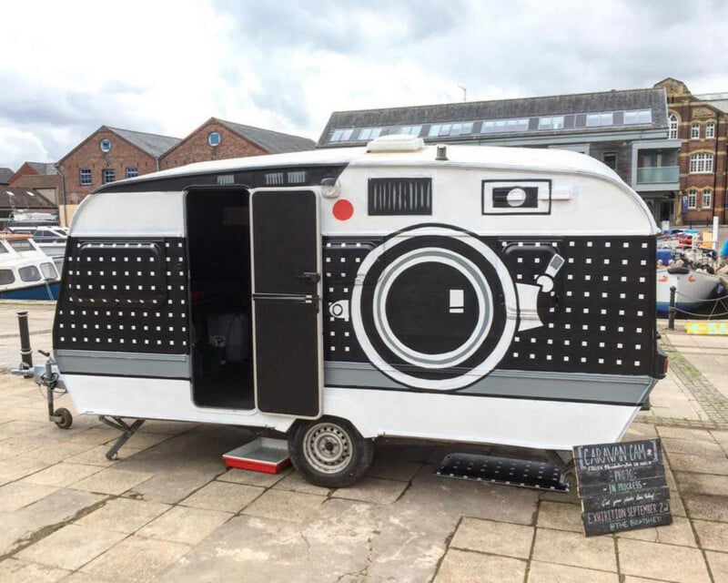 A white caravan painted to look like a retro camera is parked outdoors. The door is open, revealing a dark interior. A small chalkboard sign stands on the ground beside the caravan. Buildings are in the background.