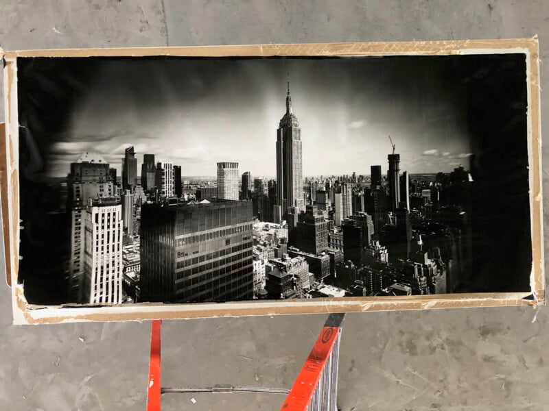 Black and white photograph of New York City’s skyline centered on the Empire State Building, displayed on a cardboard-backed frame resting atop a red ladder, viewed from above.