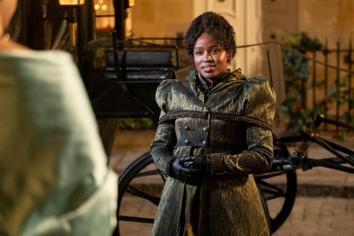 Masali Baduza as Michaela Stirling in Bridgerton standing on the road by a carriage, wearing a coat and gloves.