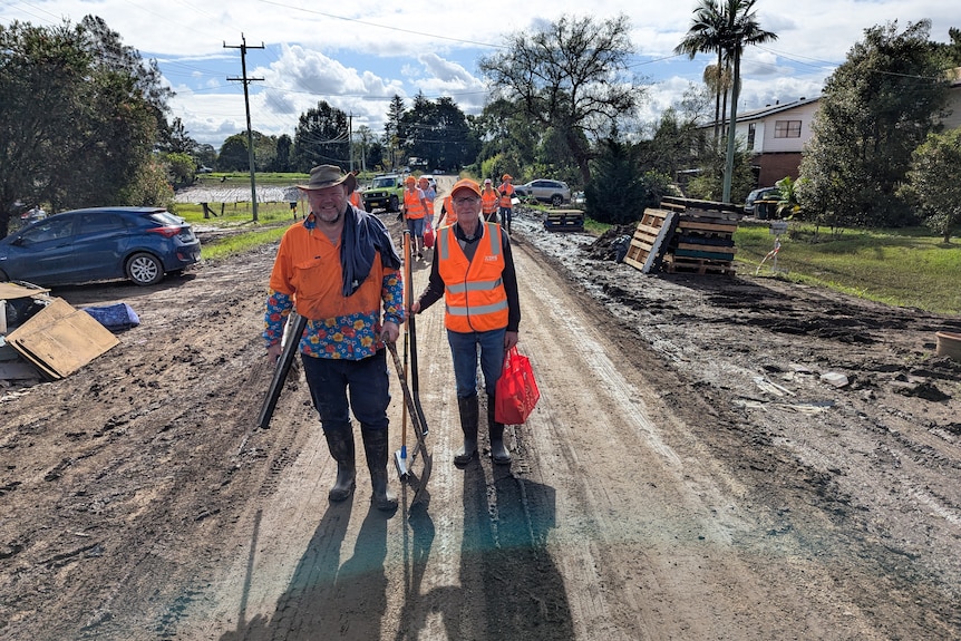 People in orange vests carry tools as they walk along a muddy street.