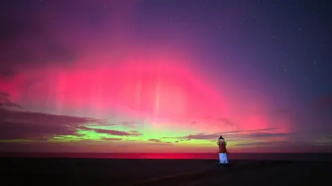 Nigel Fairclough Landscape shot of the northern lights from the Point of Ayre. A lighthouse can be seen looking over the coast whilst the sky is lit in bright pinks, greens and purple.