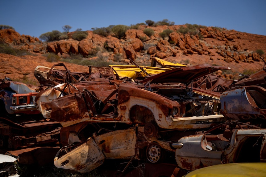 A pile of rusty cars with a hill and blue sky in the background.