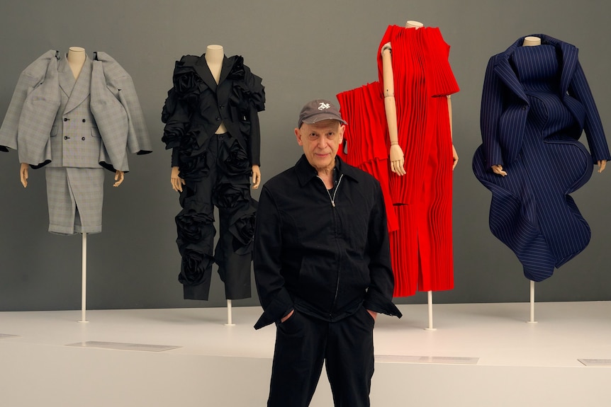 Adrian Joffe stands with his hands in his pockets at the exhibition in front of four bright mannequins in Kawakubo designs