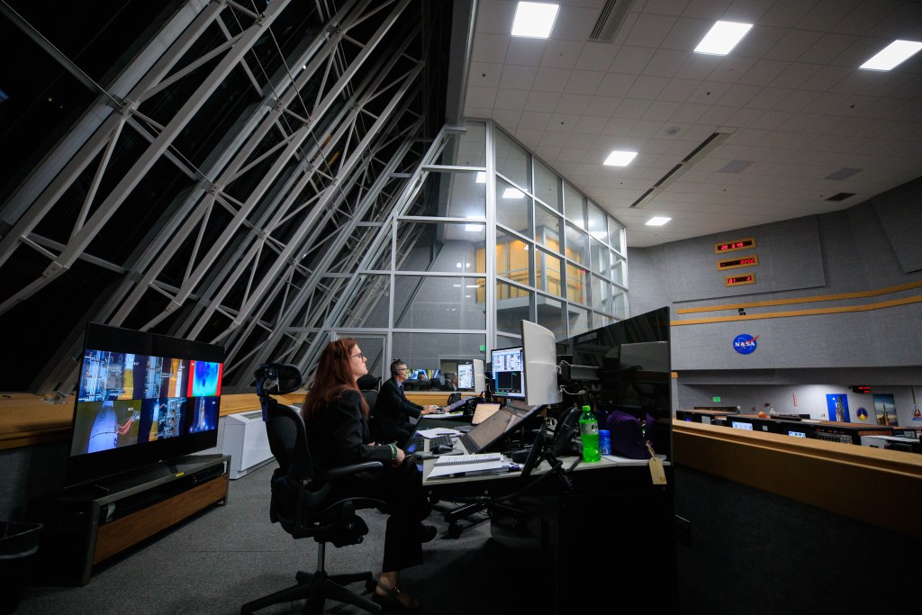 Two launch engineers--a woman with red hair and a man with short, dark hair--look intently at computer screens in an elevated platform overlooking Firing Room 1 in NASA's Launch Control Center.