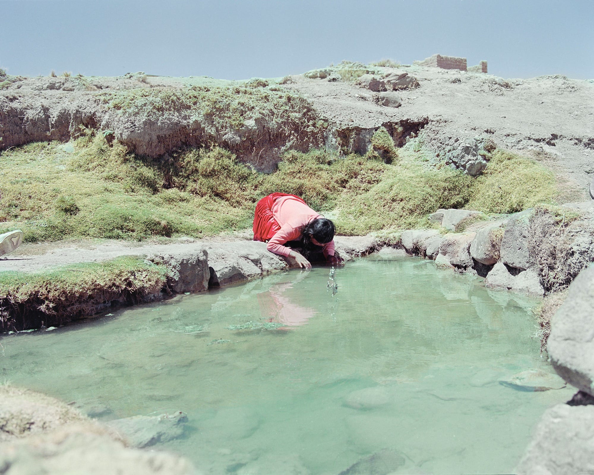 A photograph by River Claure of a figure kneeling beside a small pool in a rocky landscape