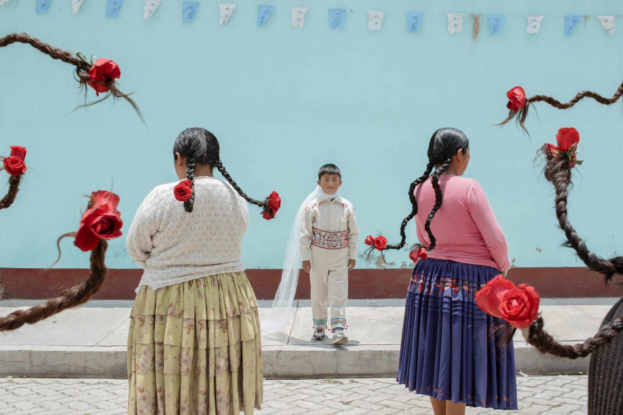 A photograph by River Claure of Bolivian women's braids and local dress, with a young boy in the center of the frame wearing white garments