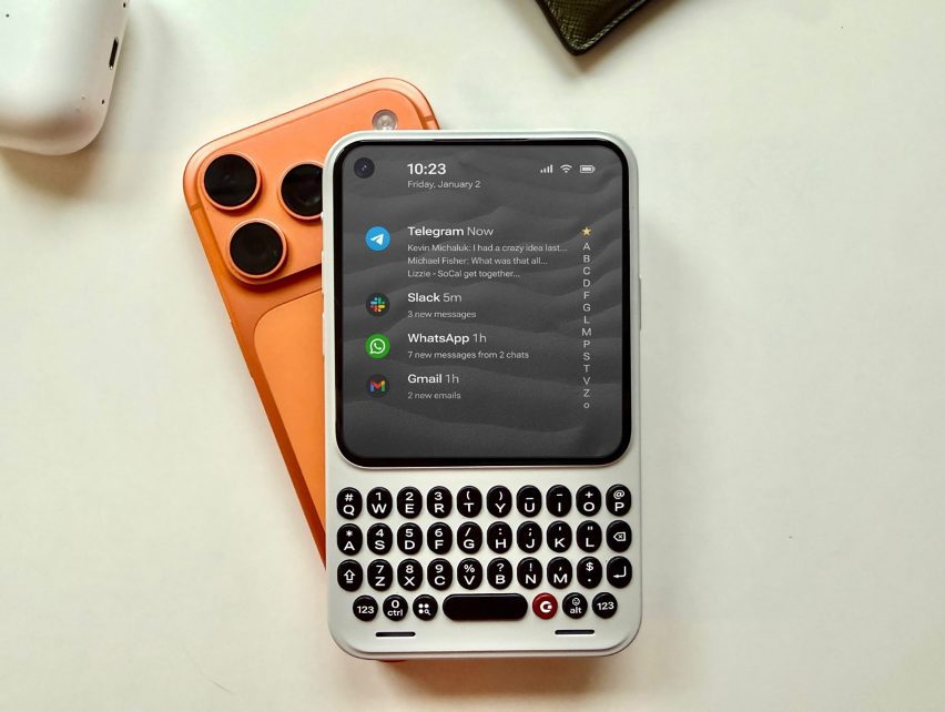 Photo of the Clicks Communicator phone lying on a desk on top of a conventional smartphone, showing its more compact size and QWERTY physical keyboard on the bottom half and a screen with messages from various apps above it