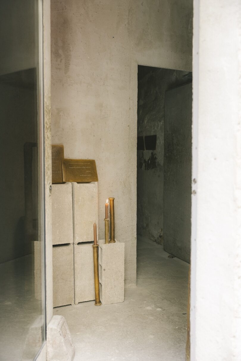 A small, plain room with stone blocks, a plaque, and several lit candles placed on the blocks and floor near the entrance.