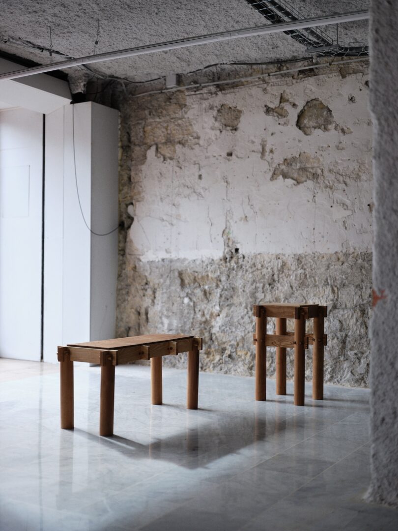 Two wooden stools with simple, geometric designs stand on a shiny floor in a room with rough, unfinished plaster walls and exposed wiring.