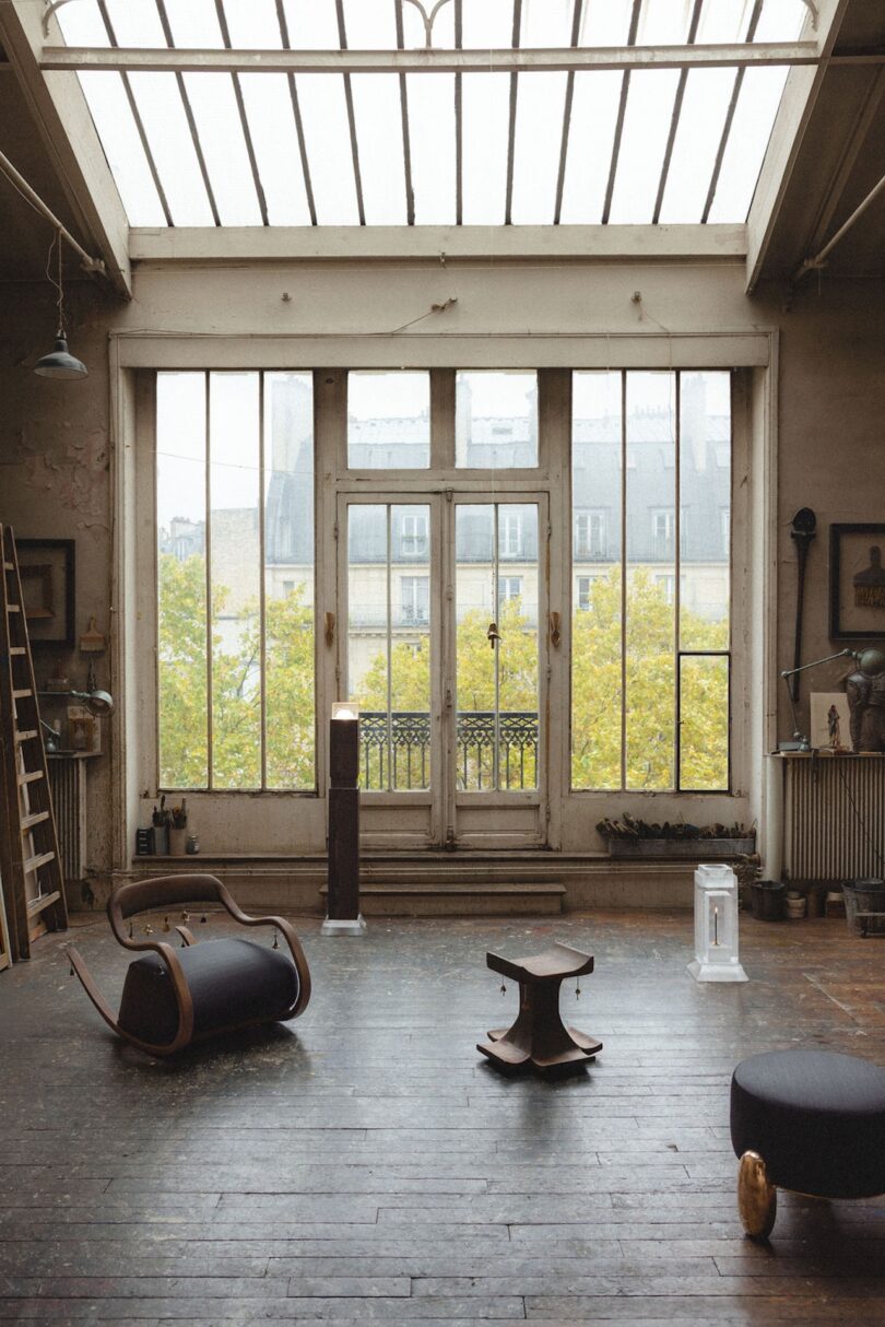 A spacious, rustic room with large skylights and tall windows, wooden floors, scattered modern furniture, and a view of trees and buildings outside.