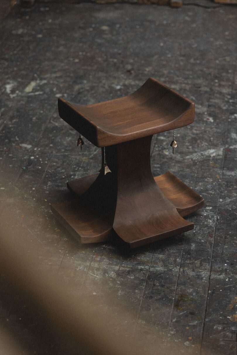 A dark wooden, sculptural stool with a curved seat and base sits on a worn, paint-splattered floor; small charms hang from the stool's edges.