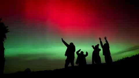 BBC WeatherWatchers/Highland Dean Silhouetted figures raise their arms toward the sky beneath a striking display of red and green aurora lighting the night.