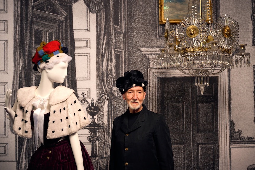 Stephen Jones stands wearing a soft black fabric crown, next to a tall mannequin wearing a tartan fabric crown