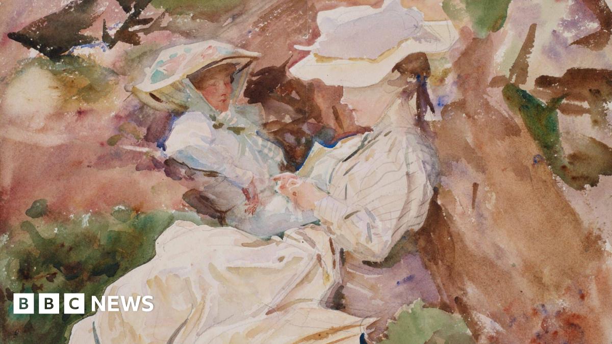 A man in a colourful red shirt with black glasses and long grey hair smiling at camera infront of artwork