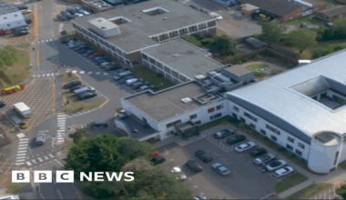 An aerial view of the large Derwent Centre complex in Harlow. It features a modern, white, curved-roof structure at the centre. Surrounding it are several older, rectangular buildings arranged along internal roads and parking areas. Cars are parked in multiple spaces and the site is bordered by trees.