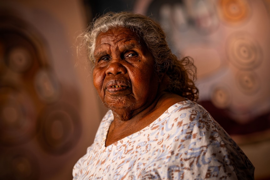 Aboriginal woman, Tuppy, has grey hair and stares at the camera lens. She is wearing a patterned top