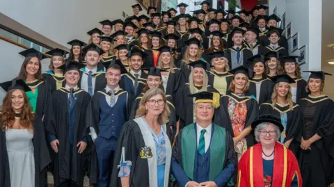 Ulster University Shows a large number of students in graduation gowns and mortar boards along with senior university staff at the front who are also wearing ceremonial outfits 