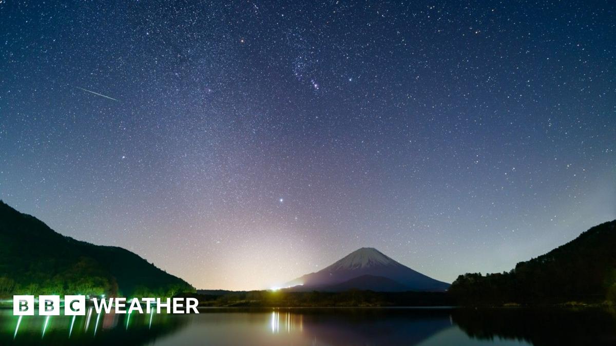 a snow capped mountain of Mount Fuji, green and yellow light reflecting onto a lake under a blue and pink starry sky with white streaks and dots or a meteor shower