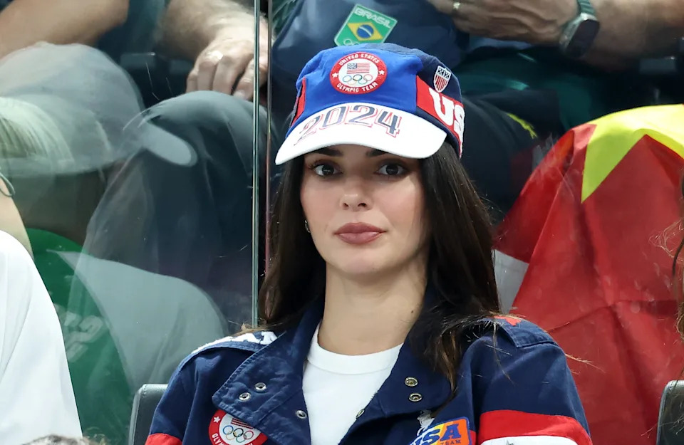 Kendall Jenner wearing a sports-themed jacket and a "2024" cap attends an event, with flags in the background