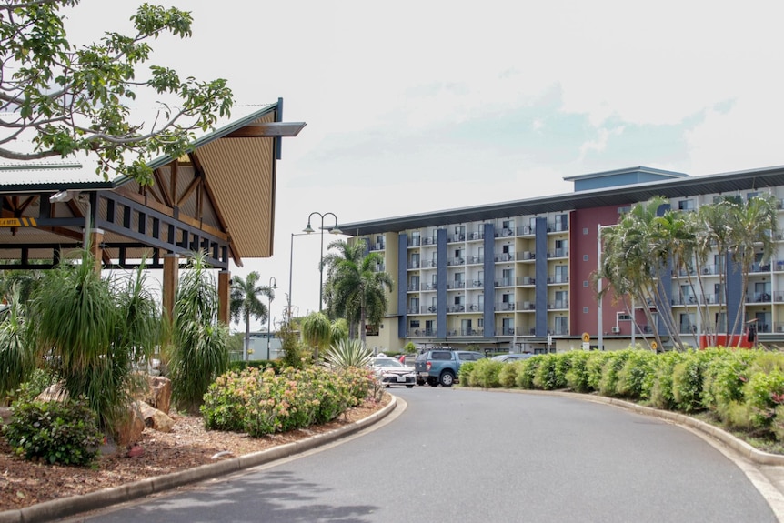 The exterior of a hotel surrounded by palm trees and greener along the entrance road