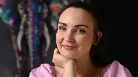 Getty Images Diona Doherty, with brown hair, tied back. She is wearing a pink top and is holding one hand up to her chin. 