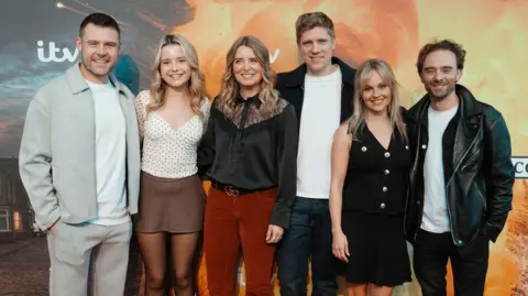 PA Media Danny Miller, Sydney Martin, Emma Atkins, Ryan Hawley, Tina O'Brien, Jack P Shepherd standing in a line and posing in front of a Corriedale backdrop with ITV branding and explosion graphics