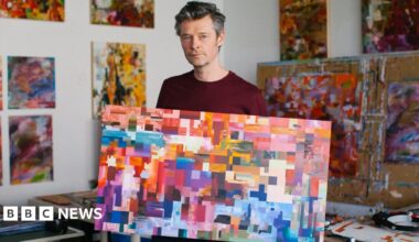 A man with grey hair in a burgundy top is standing in an art workshop with colourful abstract art in the background. He is holding a canvas in a rainbow of colours that are painted in large pixel-like squares.