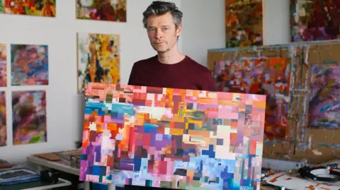 Paul Daly A man with grey hair in a burgundy top is standing in an art workshop with colourful abstract art in the background. He is holding a canvas in a rainbow of colours that are painted in large pixel-like squares.