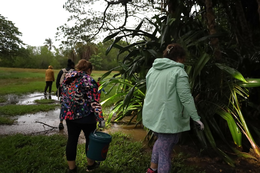 A small group of people walking through a lush public garden, in rain coats and carrying buckets.