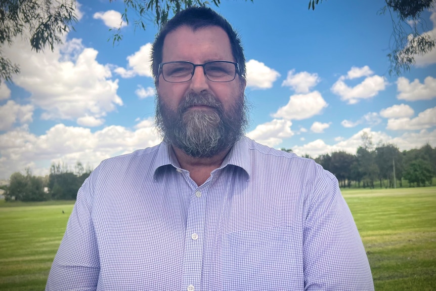Troy Stolz standing with checkered shirt and glasses, beard, brown hair, serious face