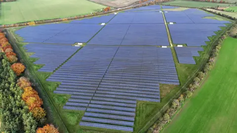 NEIL HALL/EPA-EFE/REX/Shutterstock A drone picture shows a huge solar farm near the village of Great Wilbraham, near Cambridge, from above. Thousands of panels cover a huge field. The solar farm is surrounded by fields and trees.
