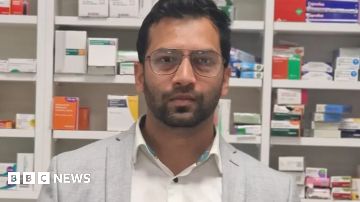 A man with a very short beard is wearing glasses, a white shirt and a grey blazer. He is standing in front of row of medication boxes.