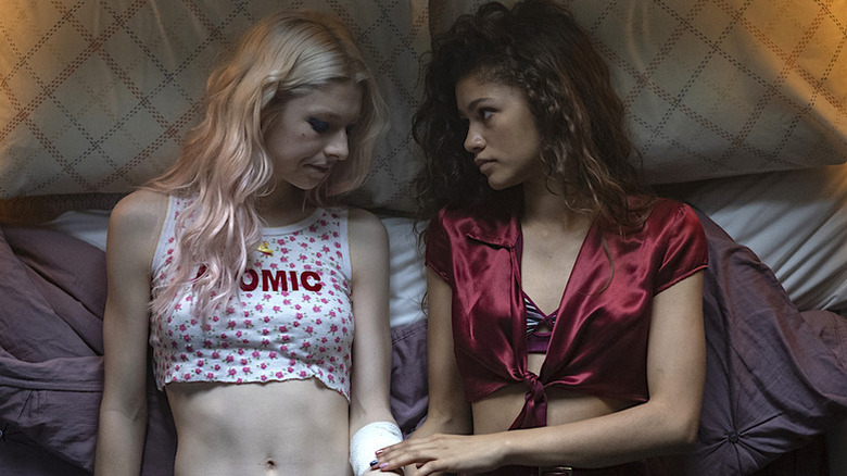 Hunter Schafer as Jules in bed with cropped white shirt next to Zendaya as Jules wearing red cropped top touching Jules' bandaged arm in Euphoria