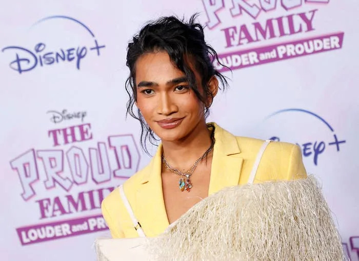 Bretman Rock in a stylish outfit with fringe detailing poses on a red carpet at an event for The Proud Family Louder and Prouder with Disney Plus logos in the background