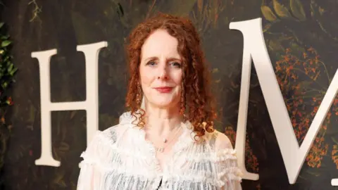 Universal/UPI Maggie O'Farrell has curly red hair which is half up. She is wearing a sheer white blouse with a black tank top underneath. Behind her can be seen a large white H and M on a green forest background. 