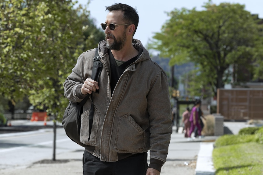 A TV still of Noah Wyle, 54, in sunglasses and motorcycle jacket, walking down a suburban street, backpack over his shoulder.