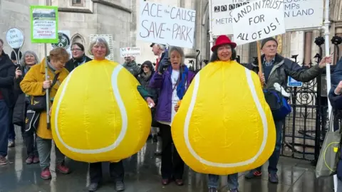 BBC London Two women dressed as tennis balls protest outside Royal Courts of Justice with other members of Save Wimbledon Park. They hold various signs with writing such as "Land In Trust For All Of Us!".