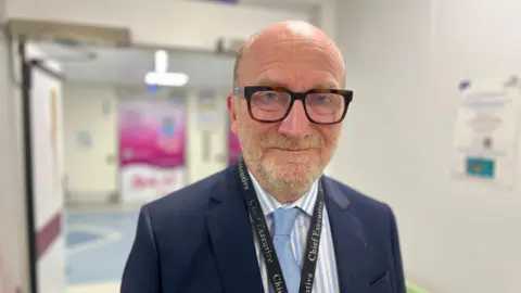 NIKKI FOX/BBC Nick Hulme stands in the corridor at Colchester Hospital. There is a blurred poster to his left and a large pink poster in the background. He is wearing a navy suit jacket and has  a lanyard around his neck that describes his position. He has a pale blue and white pinstripe shirt and a blue tie. He is wearing thick plastic black glasses and has a short beard