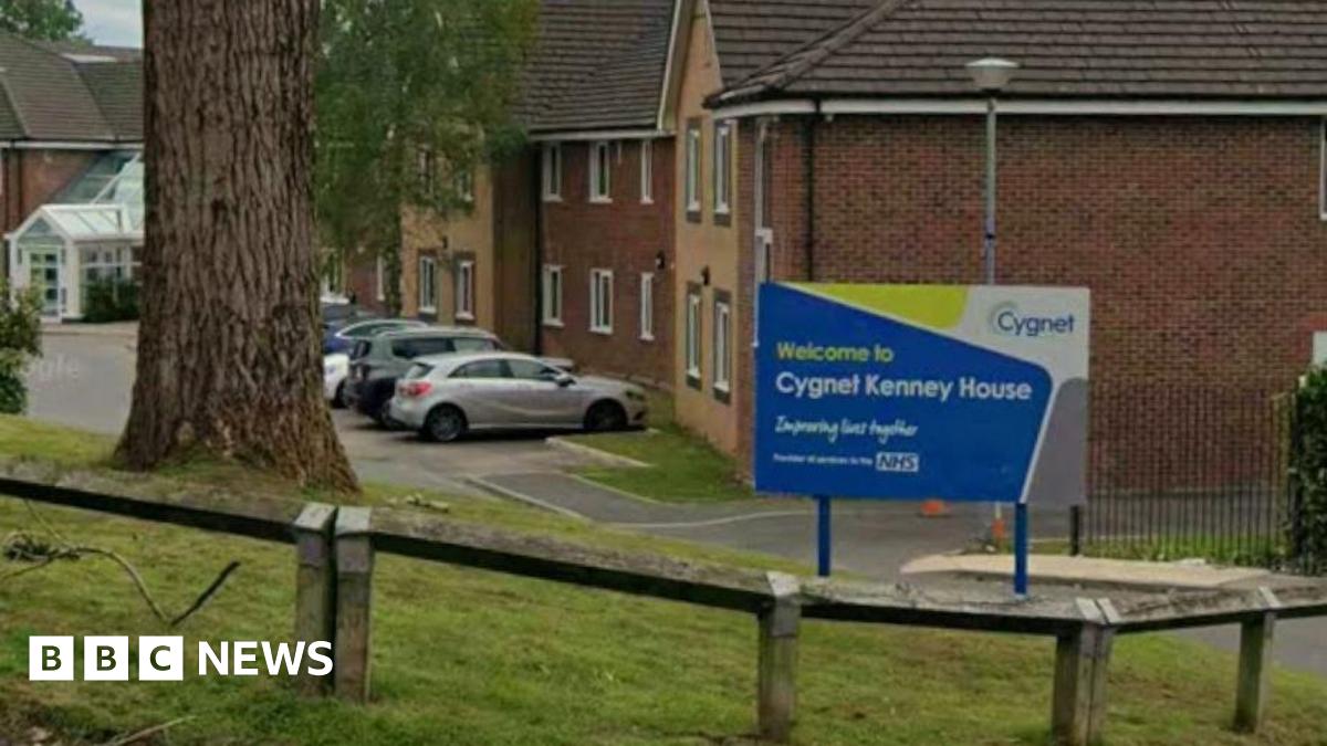 A sign for 'Cygnet Kenney House' in blue and yellow, placed on a small lawn by the entrance to a mental health unit. Cars can be seen parked in a car park while a large tree can be seen on the grass bank.