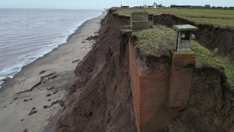 The nuclear bunker - a red brick building - is partially exposed in a muddy clifftop about 20m above a beach. Eroded muddy cliffs are seen in the background. 