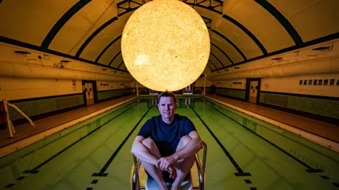 PA Media Artist Luke Jerram sat on a chair near a pool. His sculpture Helios, which is inspired by the sun, is suspended above the pool. Luke has short dark hair and is wearing shorts and a dark coloured top. His knees are bent up to his chest and his arms are folded over his legs. He is looking at the camera and smiling. 