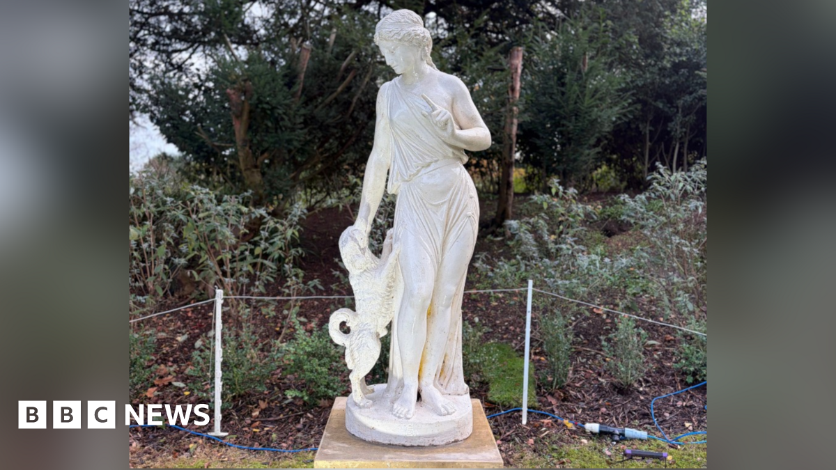 A close up of the new statue. It is white alabaster. A Greek woman stands looking down at a dog, that is on its hind legs. Its front legs lean against her hips and she pets its head with one hand. Her other arm is angled inwards with her finger wagging at the dog. Her hair is braided back and she wears a loose toga draped across one shoulder and scooped between her legs, so you can see most of her legs. Behind the statue is a roped fence and a shrub border.
