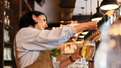 Getty Images Woman with  black, shoulder-length hair, wearing a light blue shirt and brown apron pulls a pint of lager behind the bar in a pub