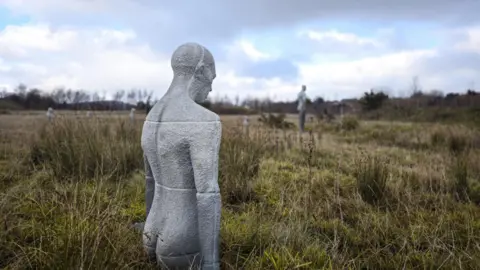 Lisa Rumsey A close view of a gray, human-shaped stone sculpture standing in tall grass, facing away from the camera. Several similar sculptures are scattered across the open field in the distance beneath a cloudy sky.