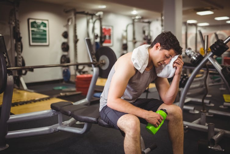 A tired man sits on a bench at the gym while dabbing his forehead with a towel.